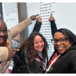 Three people are pointing to a name on a wall.
